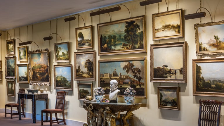 Several paintings on a cream wall, with a small table and wooden chairs in front.